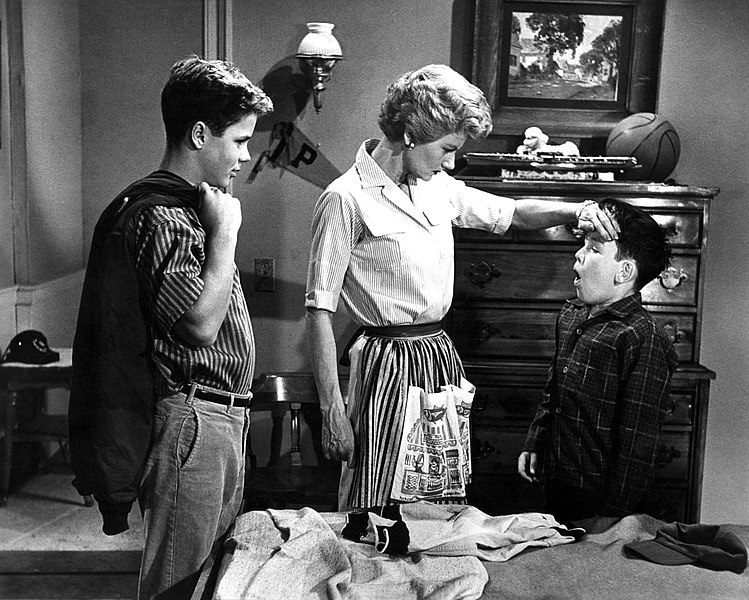 Black-and-white still from the sitcom Leave It to Beaver showing the mother character with two boys in a bedroom, placing her hand on one boy's forehead