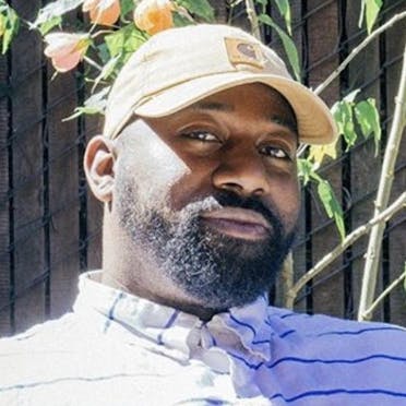Photo portrait of author Carvell Wallace wearing a tan baseball cap and white shirt with blue stripes outdoors