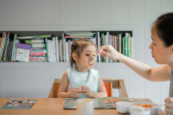 Color photo of woman feeding young child at table with spoon