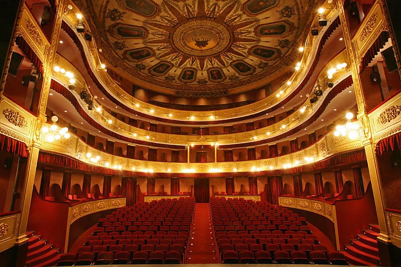 View of the interior of the Teatre Municipal in Girona, Spain showing red seats and carpet, three levels of balconies, and ornate ceiling decorations