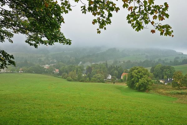 Color photo showing a view overlooking the mountain town of Sokołowsko (Görbersdorf), with green slopes, trees, and fog visible