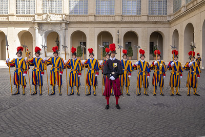 Photo of members of the Swiss Guard, in blue-and-orange striped uniforms, standing at attention outdoors in Vatican city